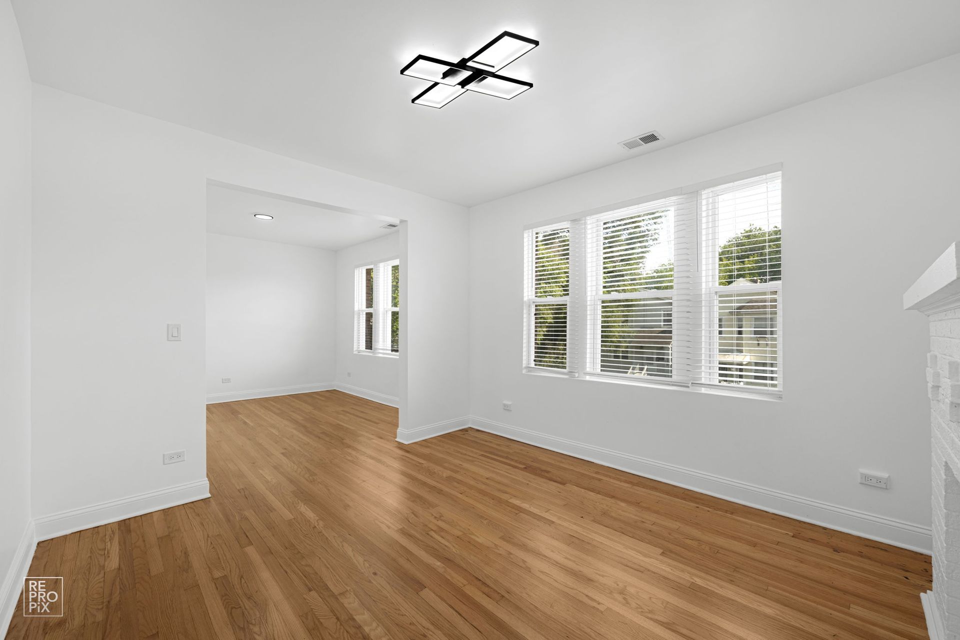 Empty room with hardwood floors, white walls, and large windows. Modern light fixture on ceiling.