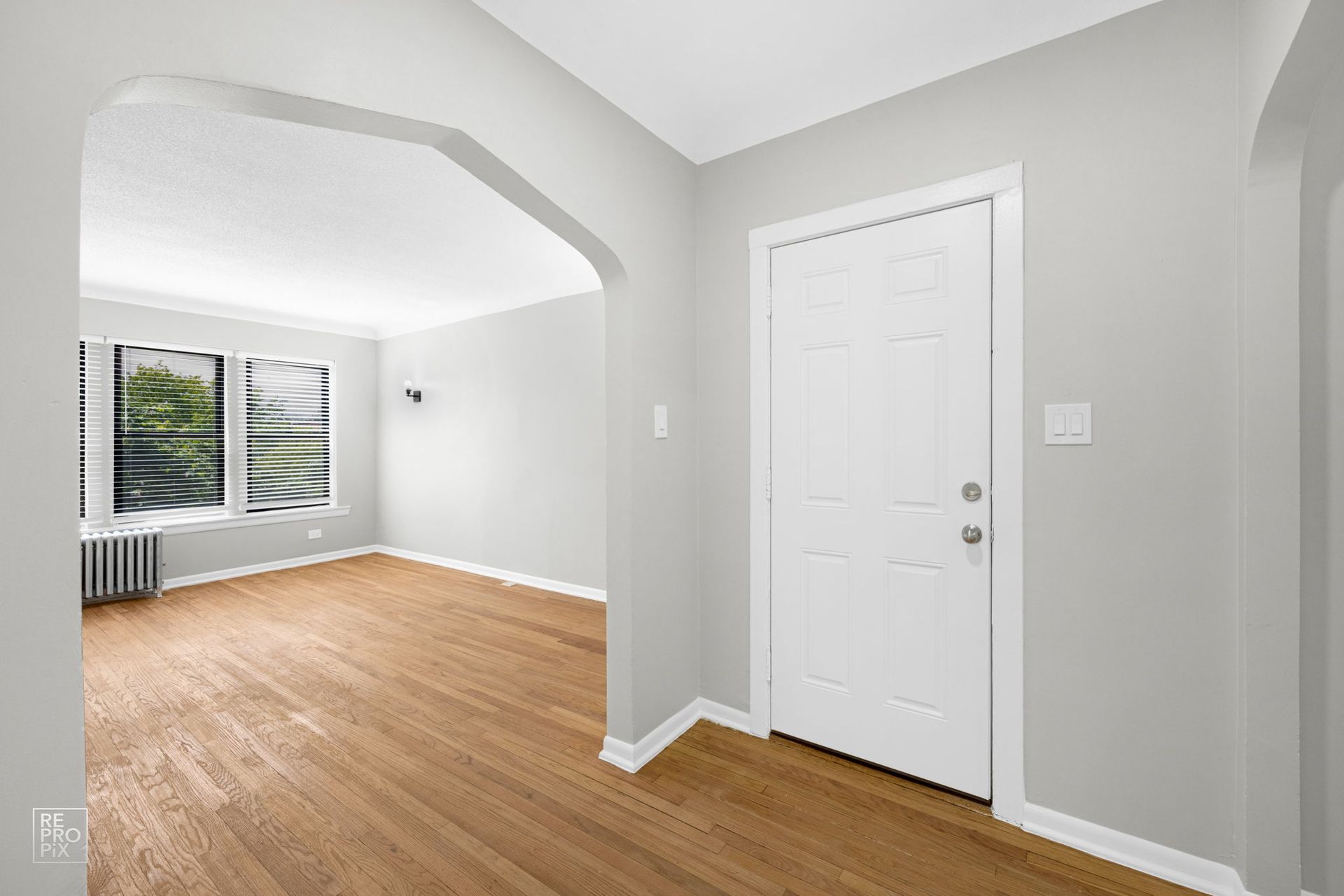 Empty apartment entryway with hardwood floors, white door, and arched doorway to a bright living room.