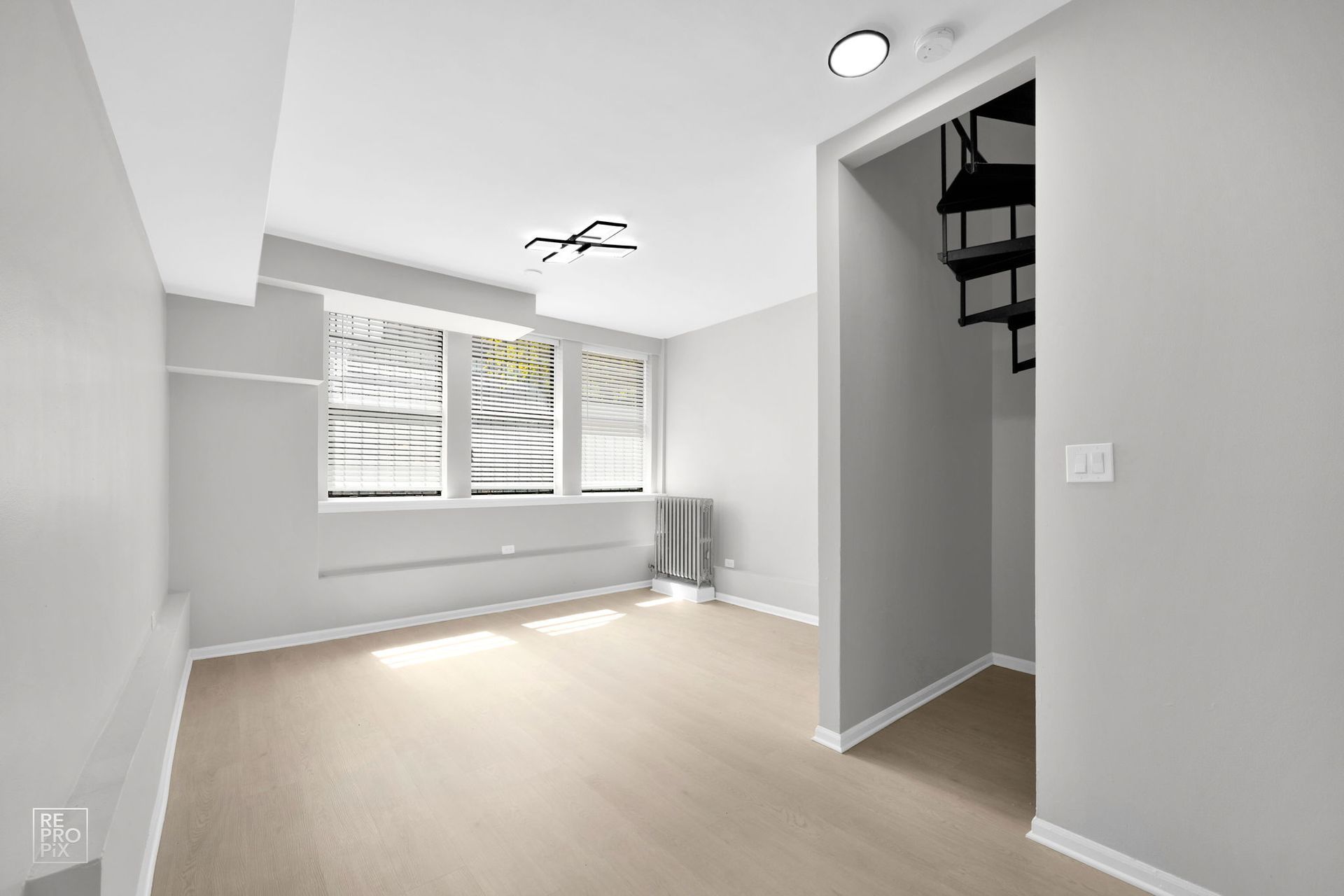 Empty room with light-colored flooring, grey walls, large window with blinds, and a black spiral staircase.