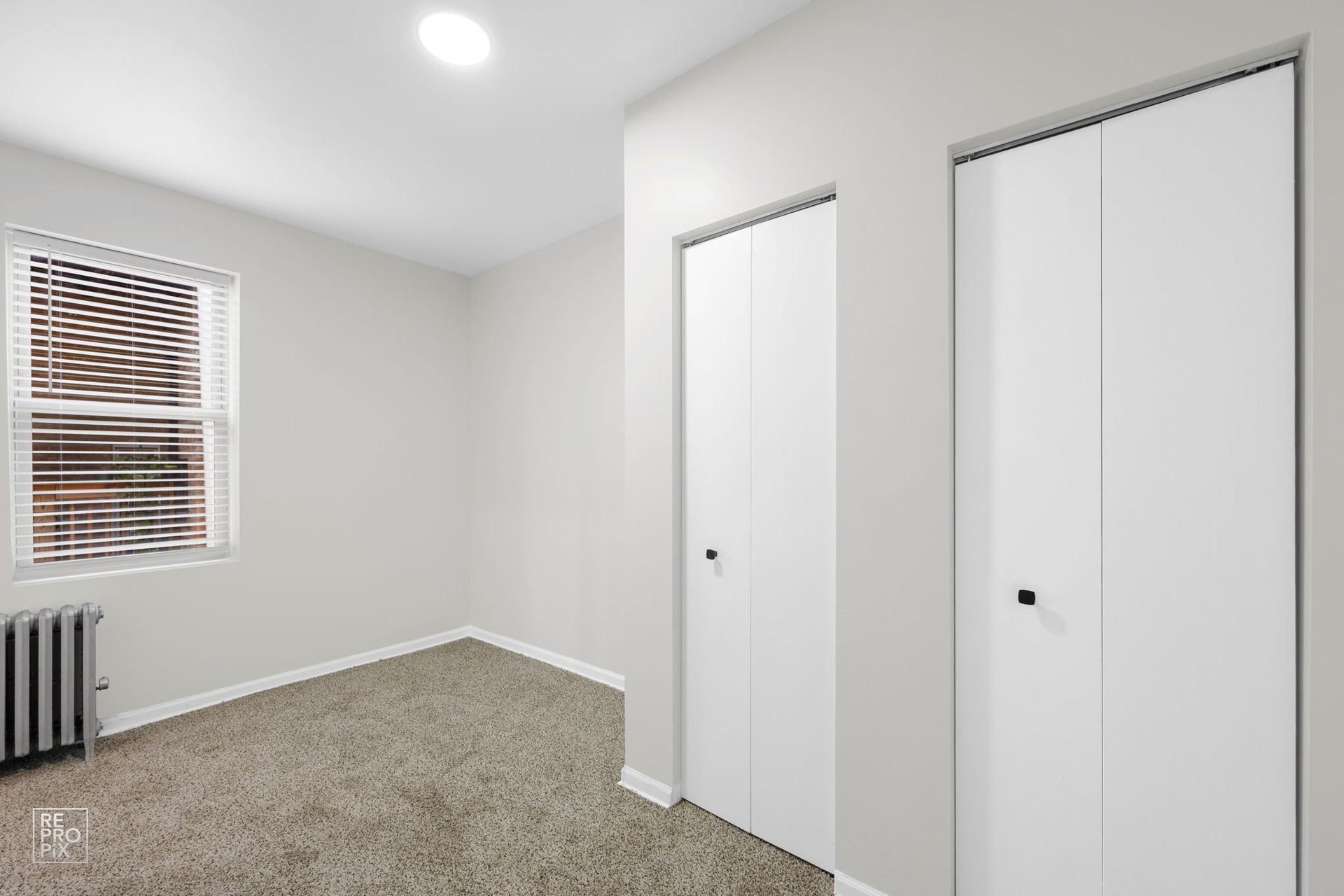 Empty bedroom with two white closet doors, window with blinds, and carpeted floor.