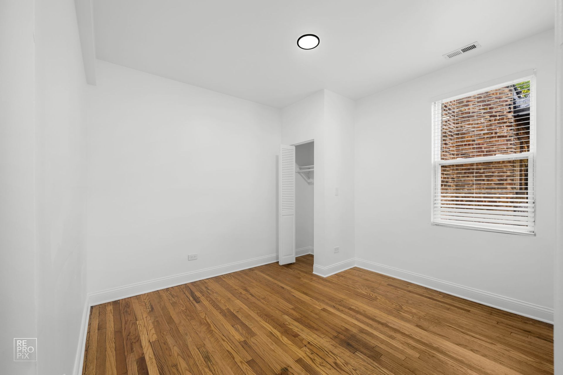 Empty room with hardwood floors, white walls, closet, and a window with brick view.