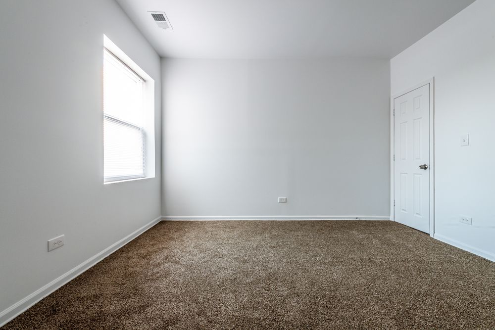 Empty room with brown carpet, white walls, a window, and a closed white door.