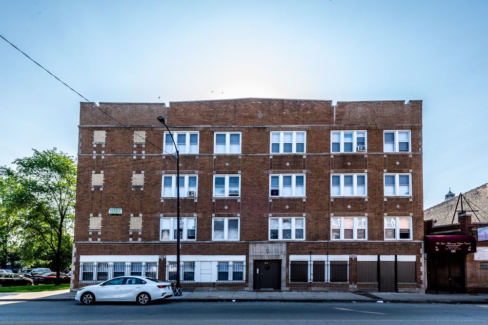 Three-story brick apartment building with white-framed windows on a sunny day. A white car parked in front.