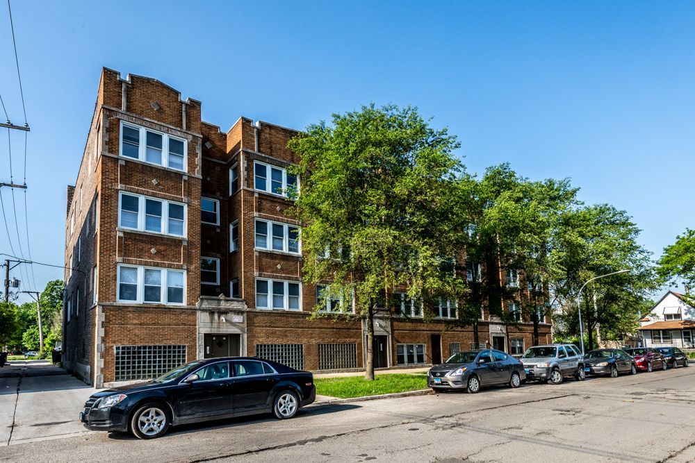 Multi-story brick apartment building on a sunny street, cars parked out front, trees in the yard, blue sky.