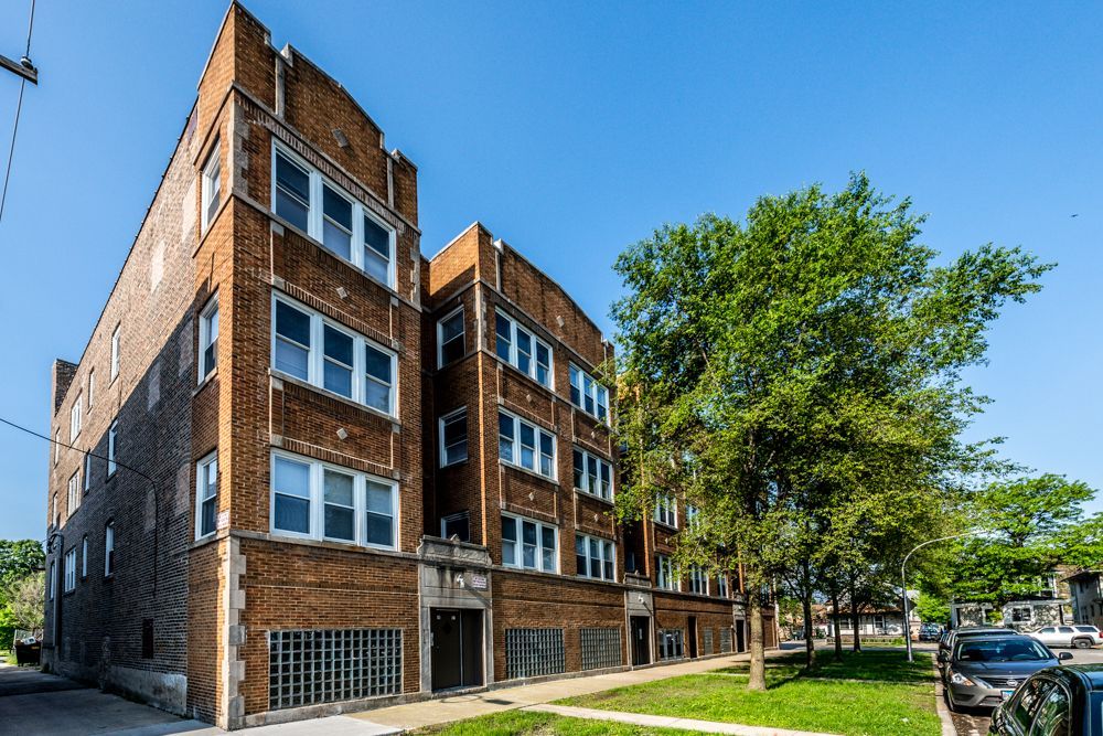 Brick apartment building with multiple windows under a blue sky, with a tree in front.