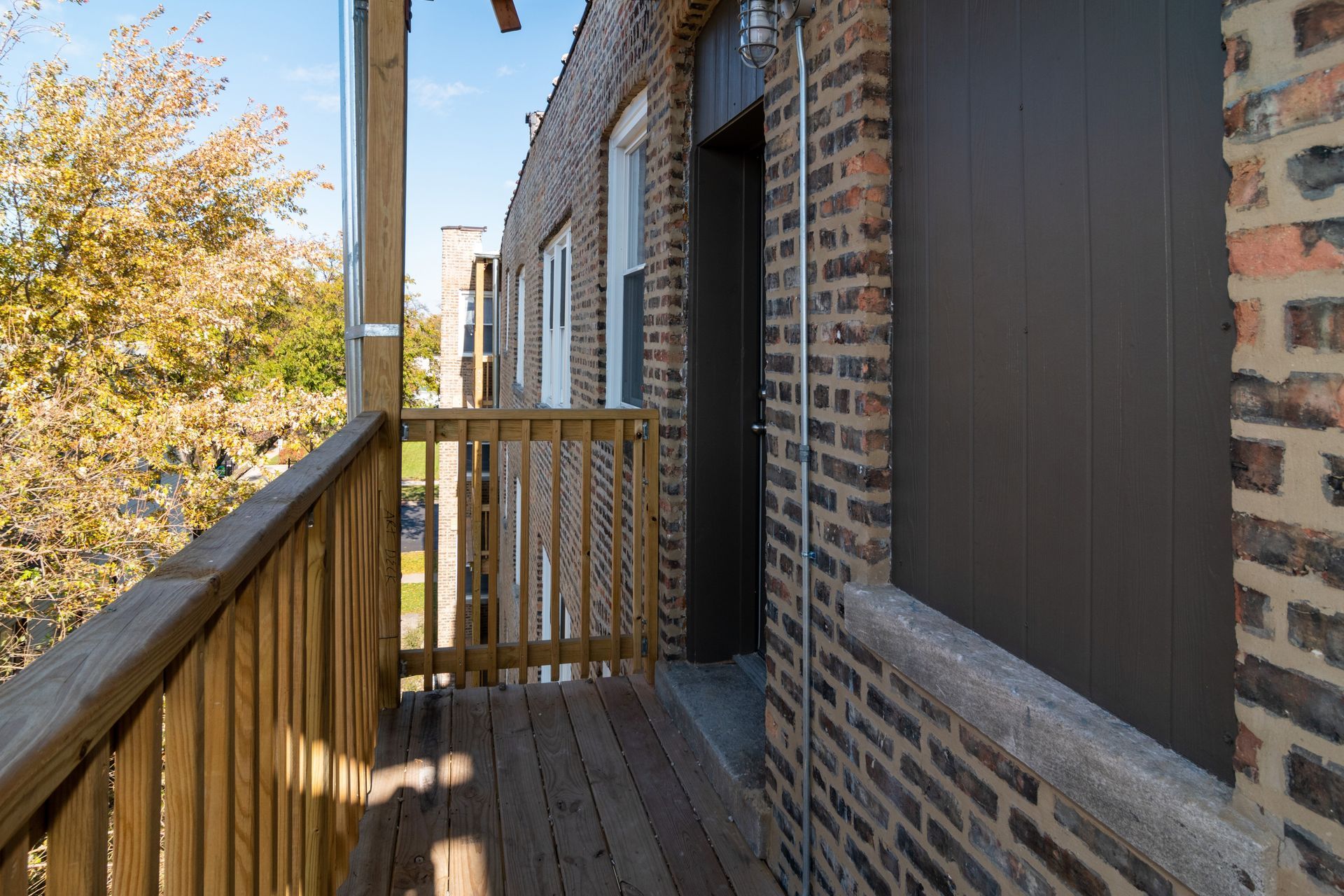 Wooden balcony attached to a brick building, with a door and trees in the background.