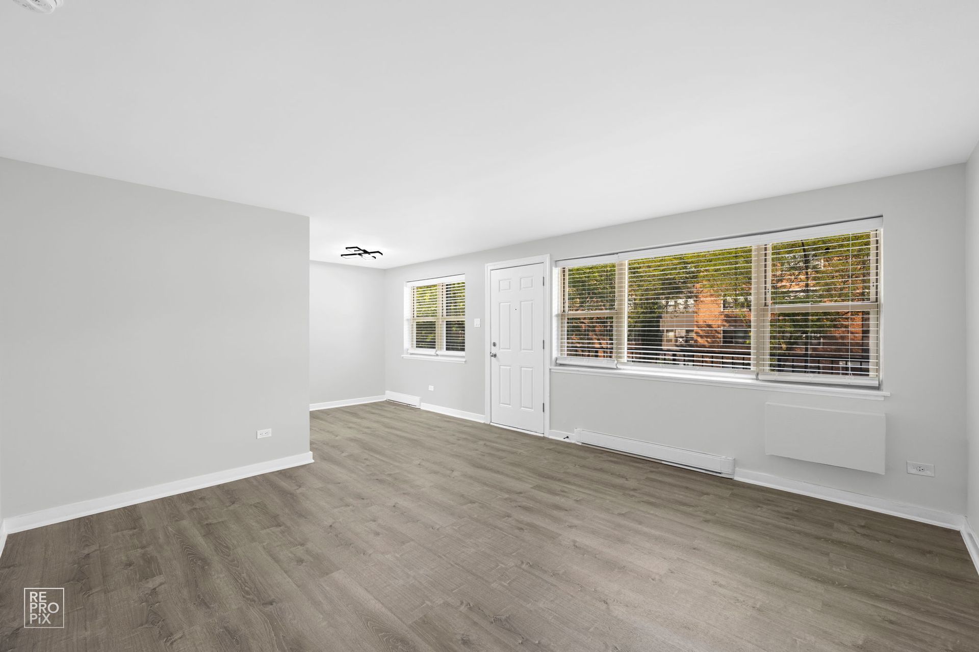 Empty living room with large windows, light gray walls, and wood-look flooring.