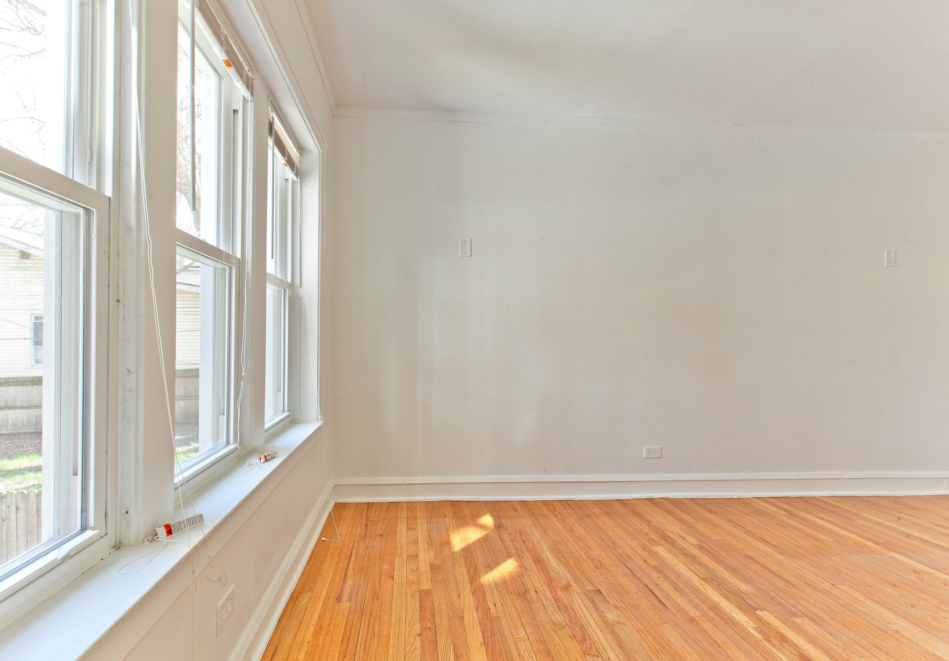 Bright, empty room with hardwood floors and white walls. Windows along one side.