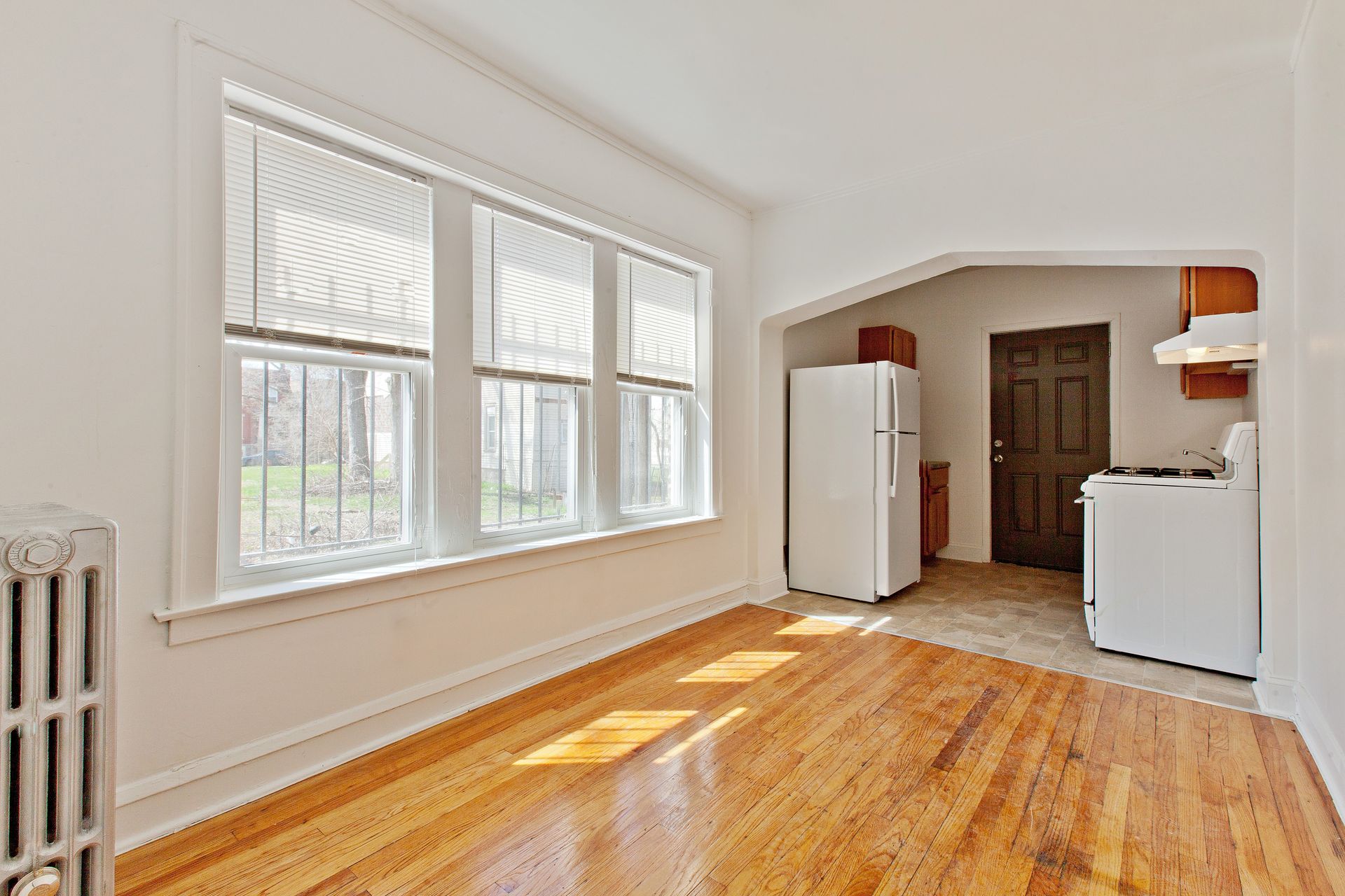 Empty sunlit apartment with wood floors, large windows, and an open kitchen.