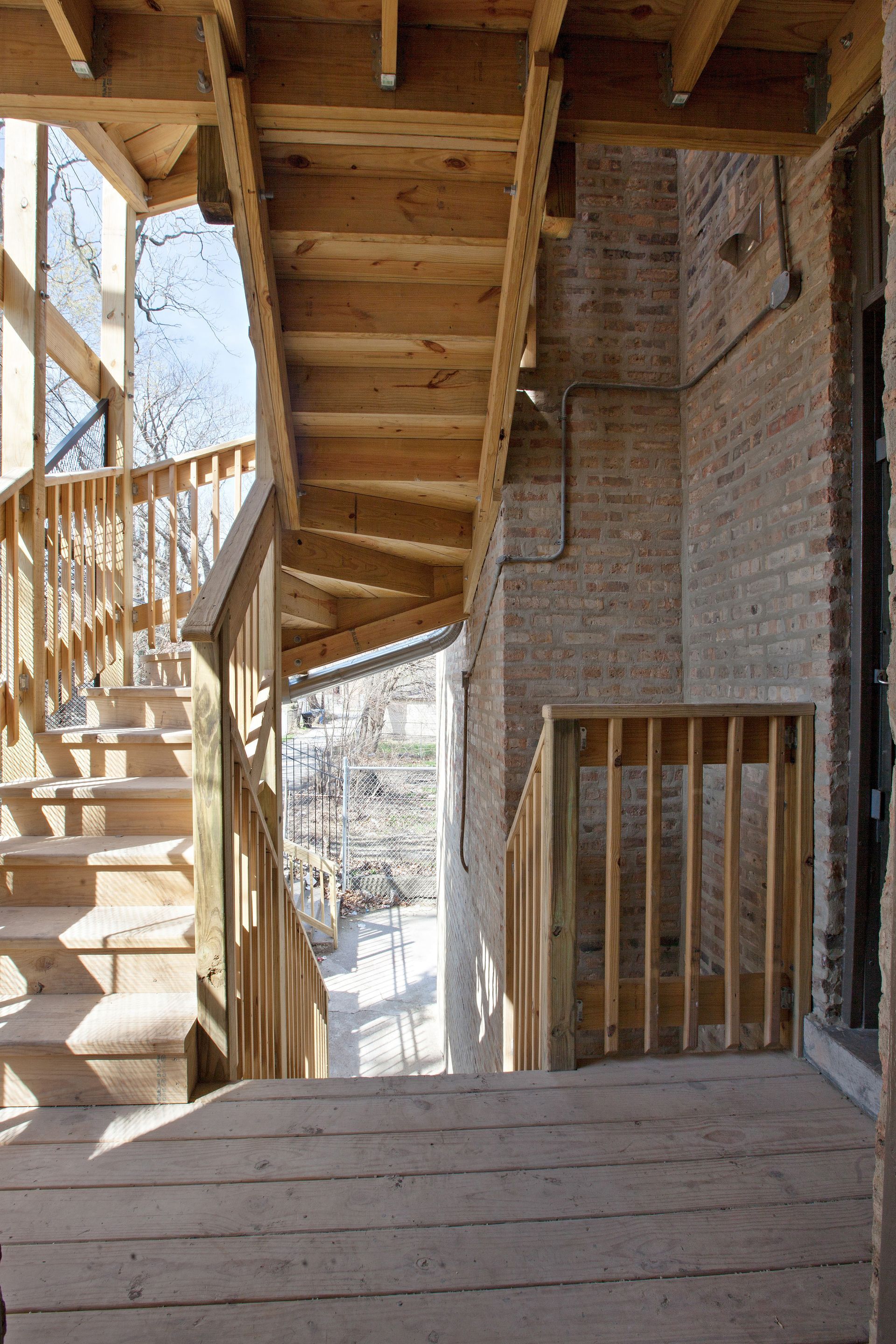 Wooden outdoor staircase and deck against a brick wall, sunlight shines in the background.