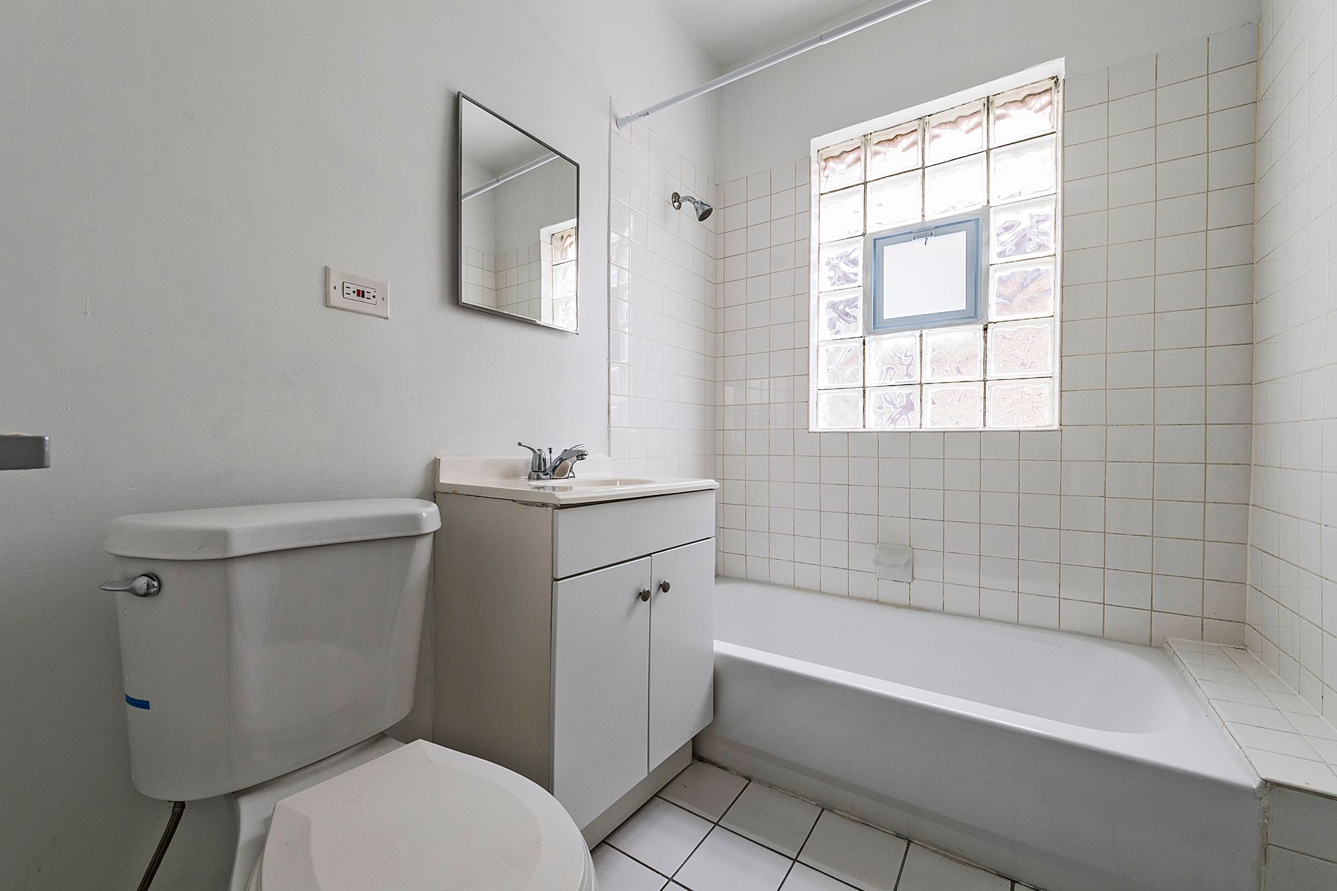 A small, white bathroom with a toilet, vanity, tub, and glass block window.