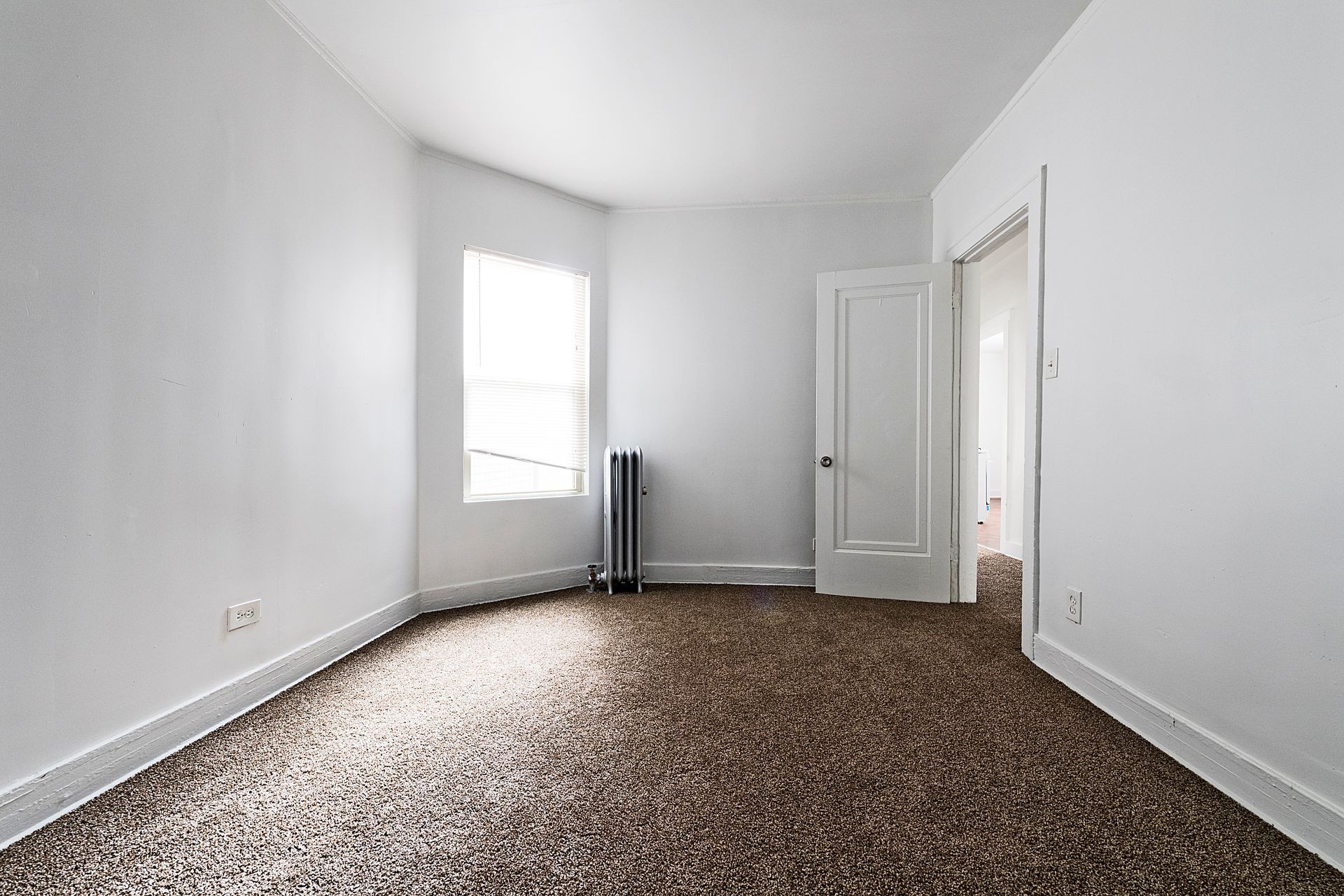 Empty room with white walls, window, and door. Brown patterned carpet, radiator.