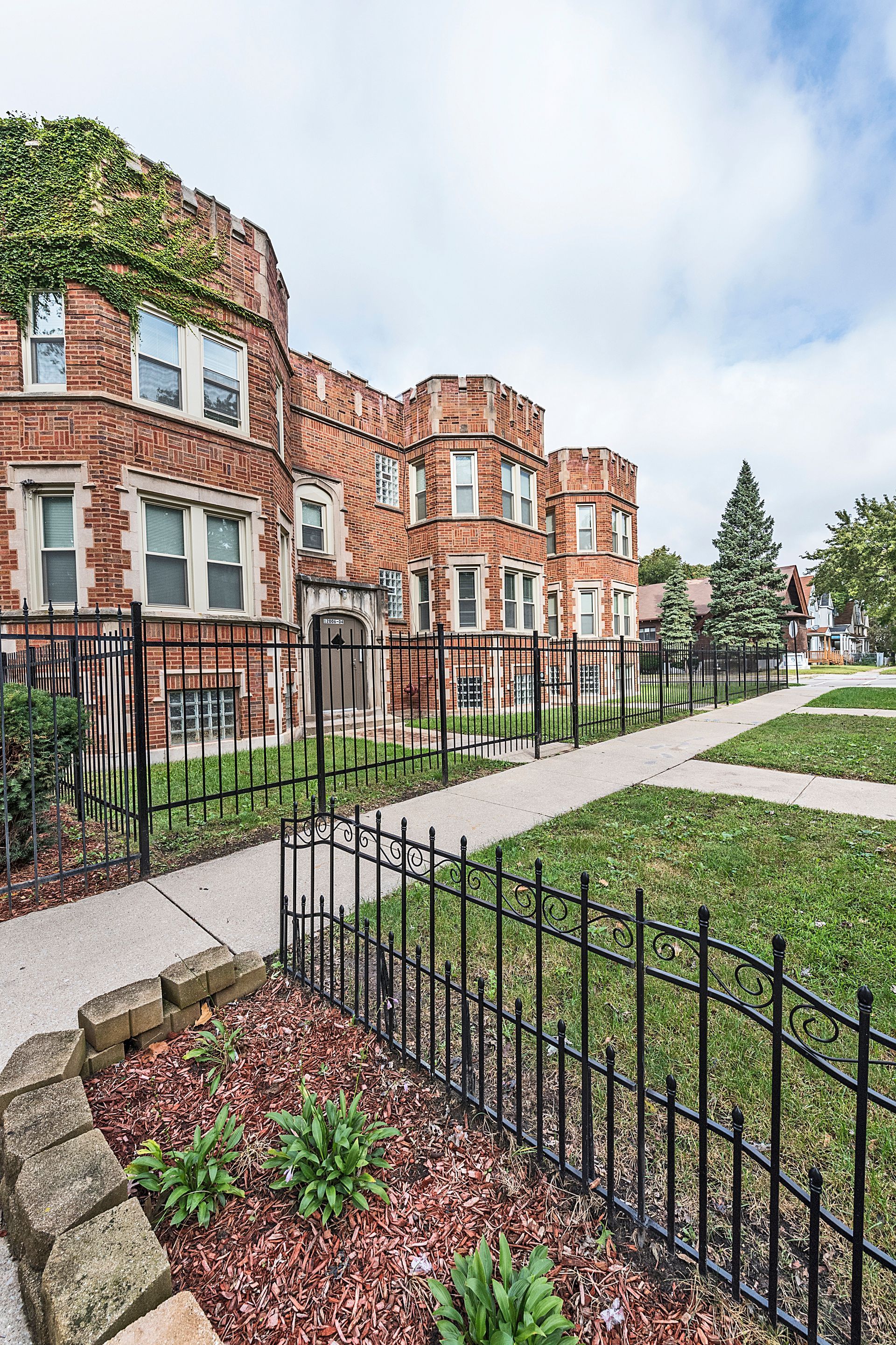 Brick apartment building with decorative turrets and wrought iron fence, sunny day.