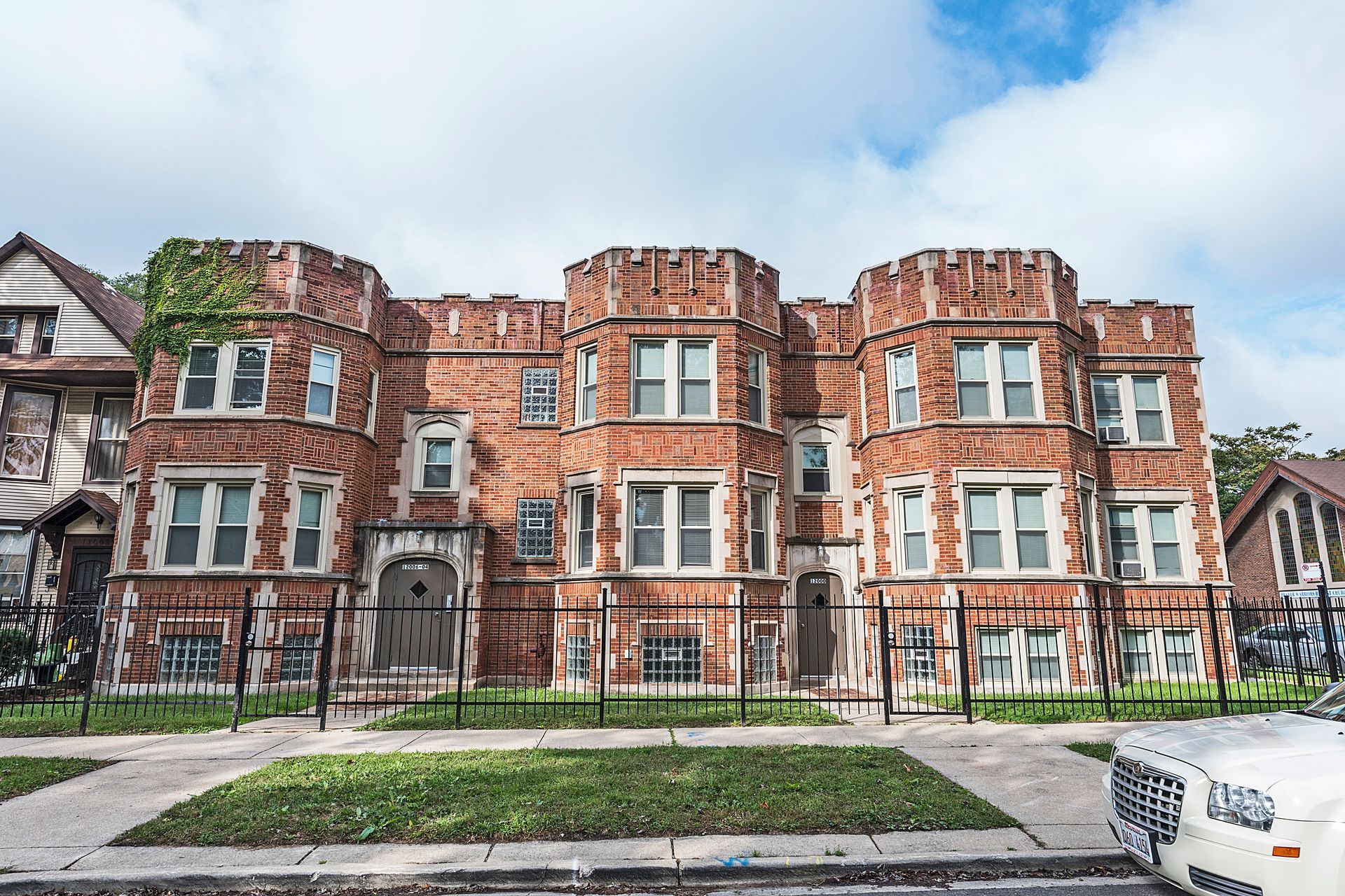 Three-story brick apartment building with decorative turrets behind a black metal fence and small lawn.