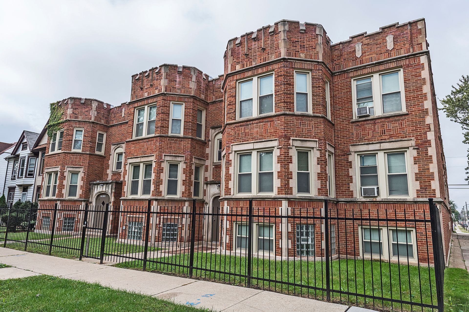 Red brick apartment building with decorative turrets and windows, behind a black metal fence on a cloudy day.