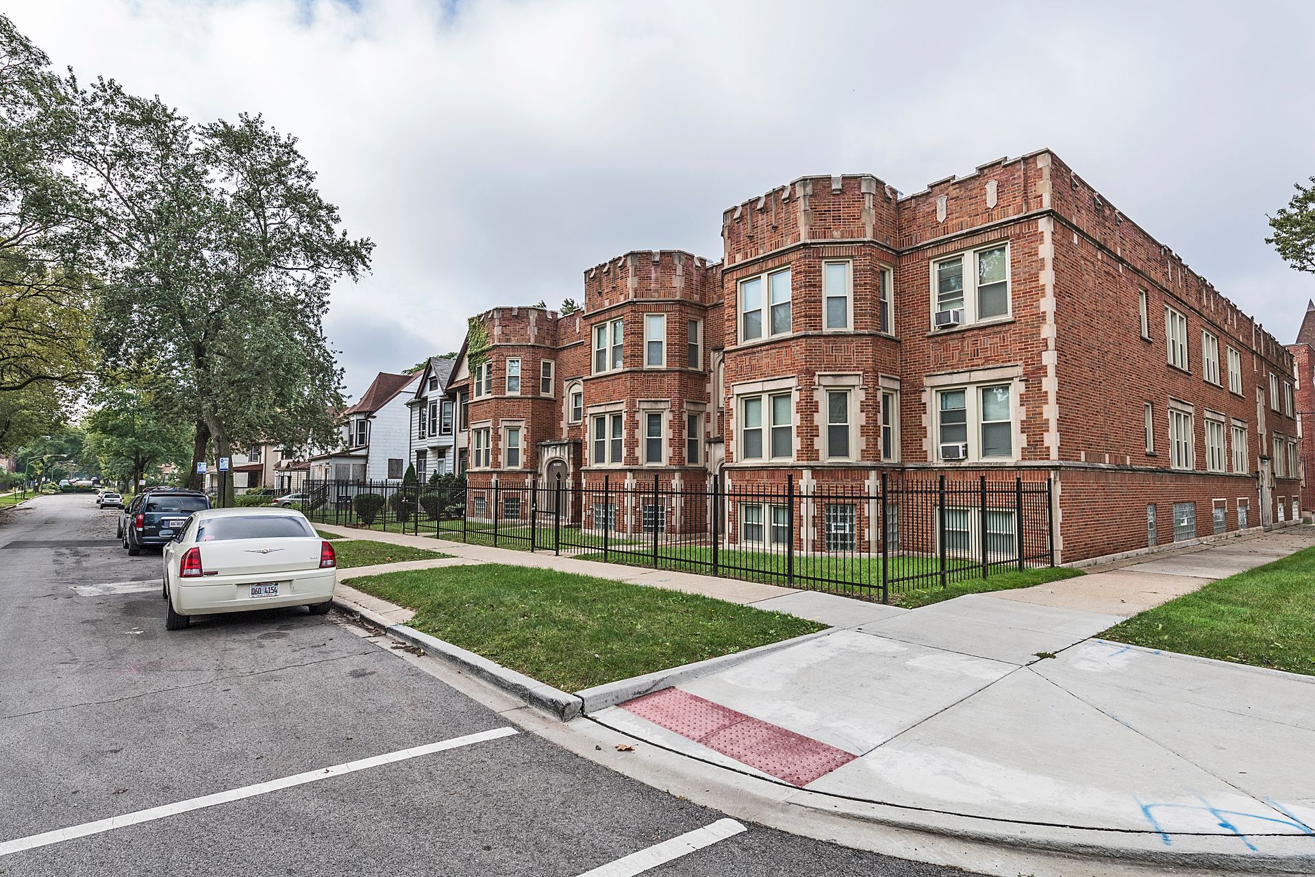 Brick apartment buildings on a street corner with parked cars, green lawns, and a cloudy sky.