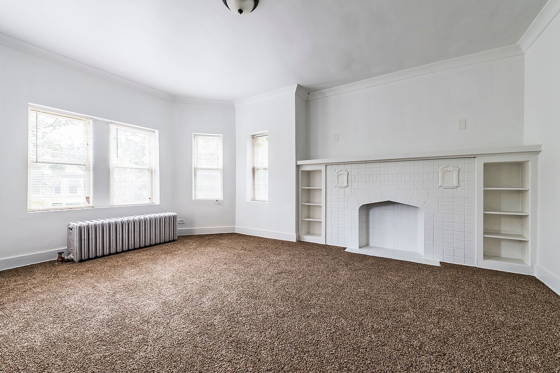 Empty living room with white walls, fireplace, and brown carpet. Windows and radiator visible.