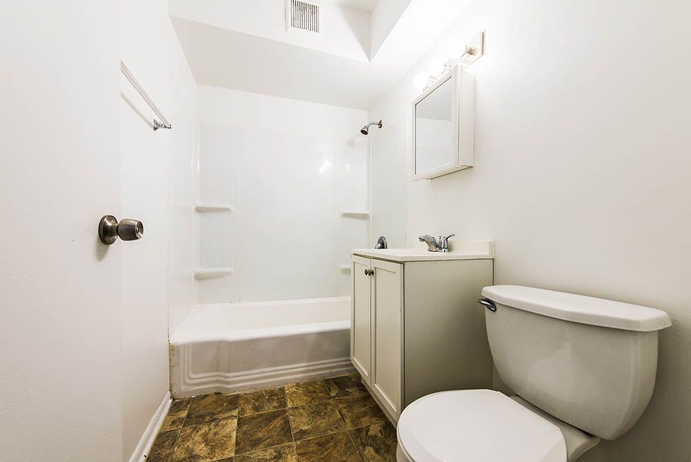 Bathroom with white walls, a bathtub/shower combo, toilet, vanity, and brown patterned floor.