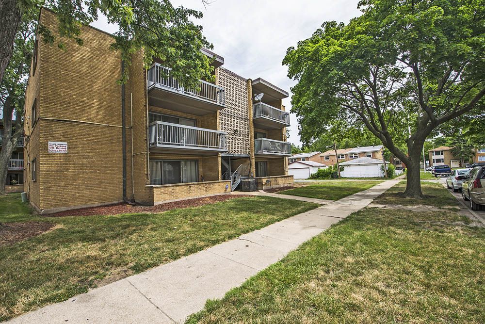 Two-story brick apartment building with balconies, sidewalk, grass lawn, and trees.