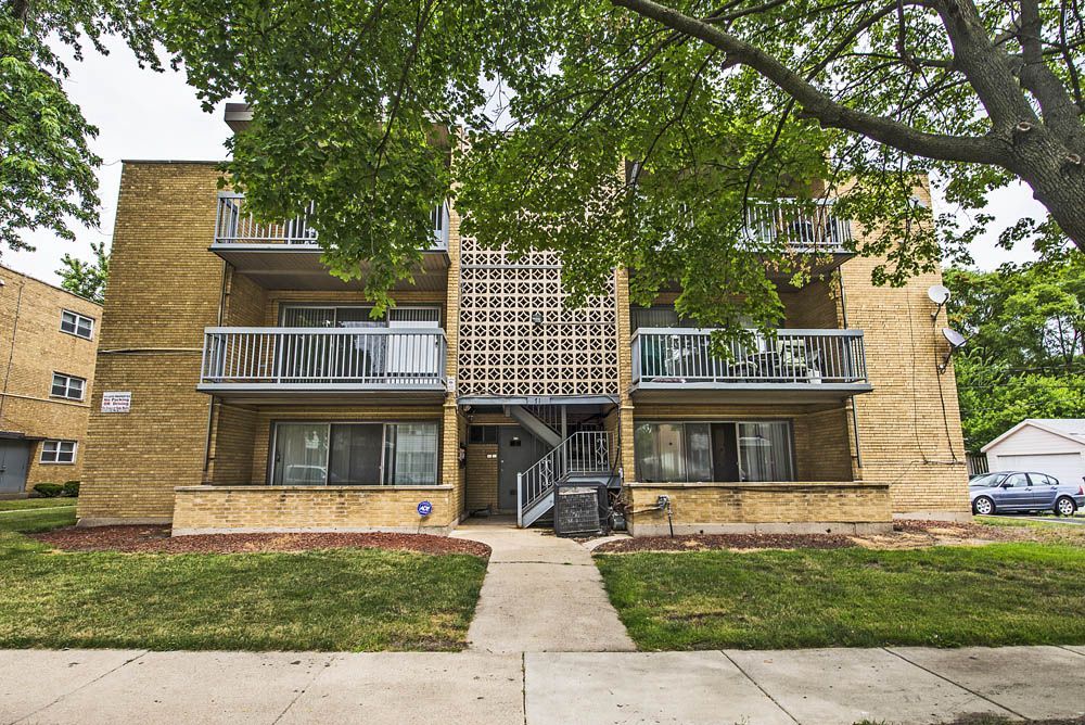 Three-story apartment building with balconies, brick exterior, and a central walkway. Green lawn and trees surround.