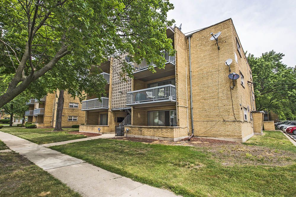 Three-story brick apartment building with balconies, trees, and green grass.
