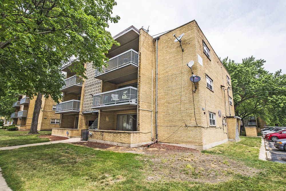 Three-story yellow brick apartment building with balconies, grass, and trees.