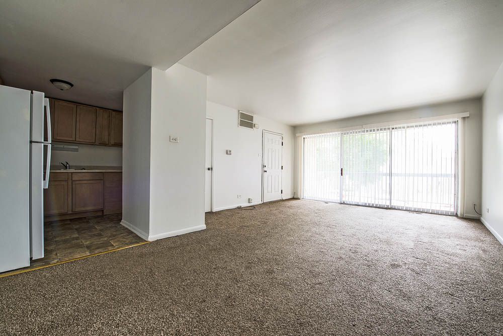 Empty apartment living room with carpet, kitchen, and sliding glass doors.