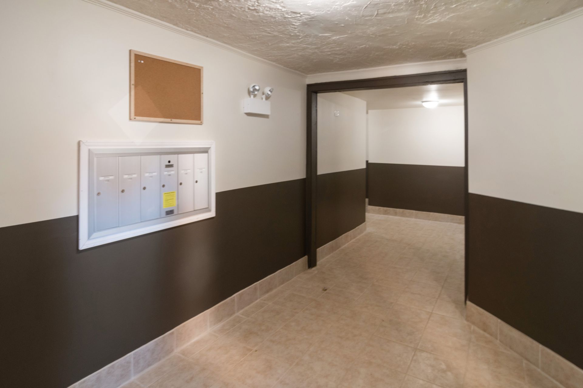 Hallway with mailboxes and bulletin board; white and brown walls, tiled floor.
