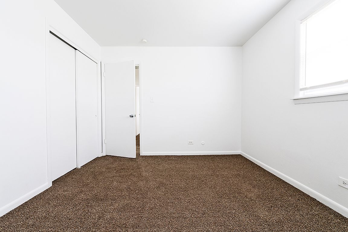 Empty bedroom with white walls, brown carpet, closed closet doors, and open door.