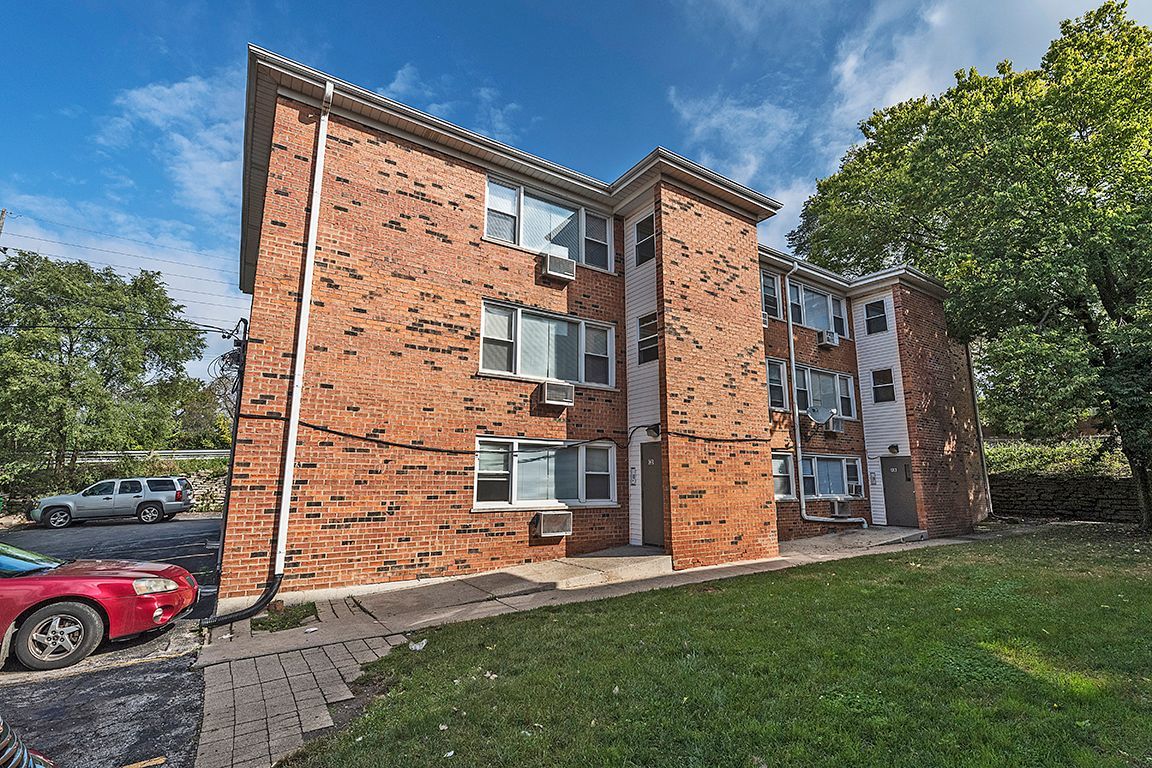 Red brick apartment building with white window frames, on a grassy hill with parked cars.