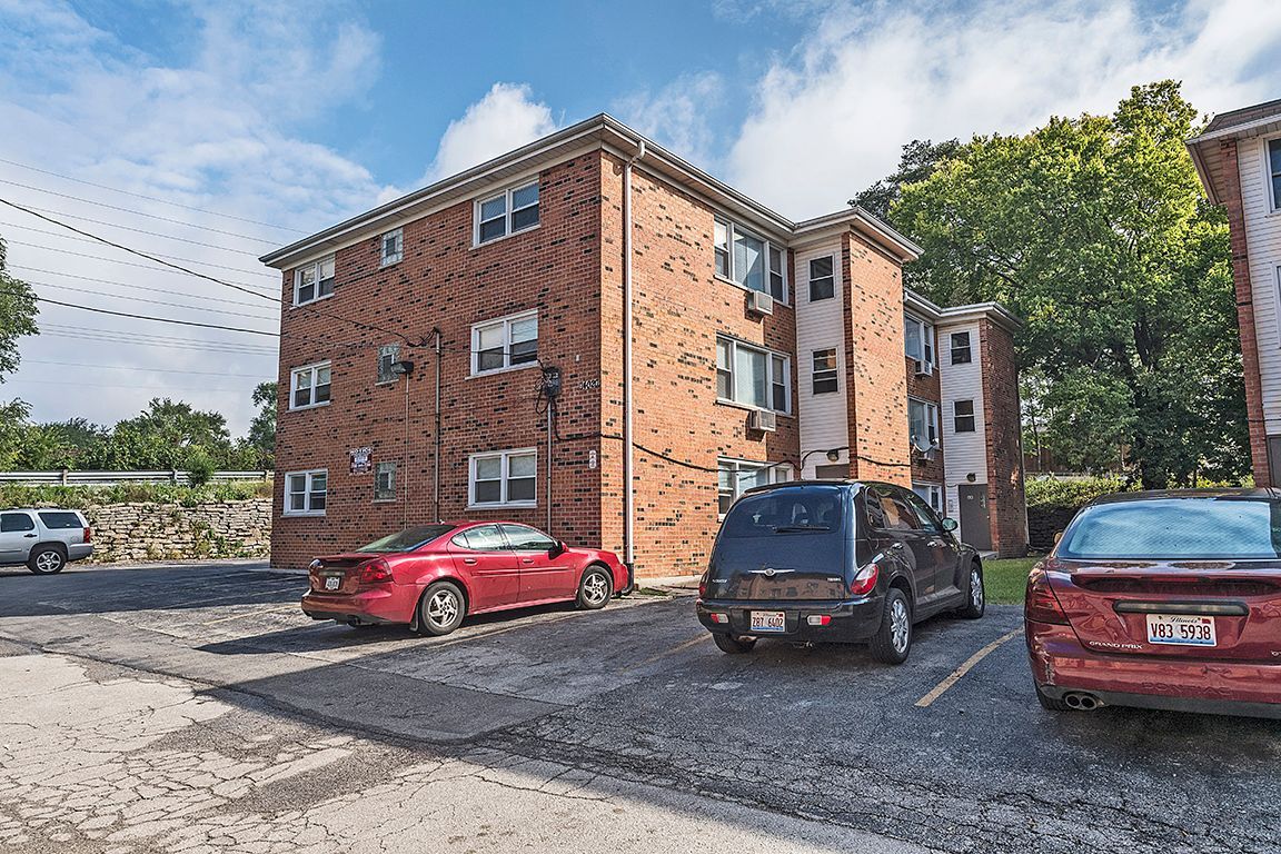 Brick apartment building with parked cars in a gravel lot under a partly cloudy sky.
