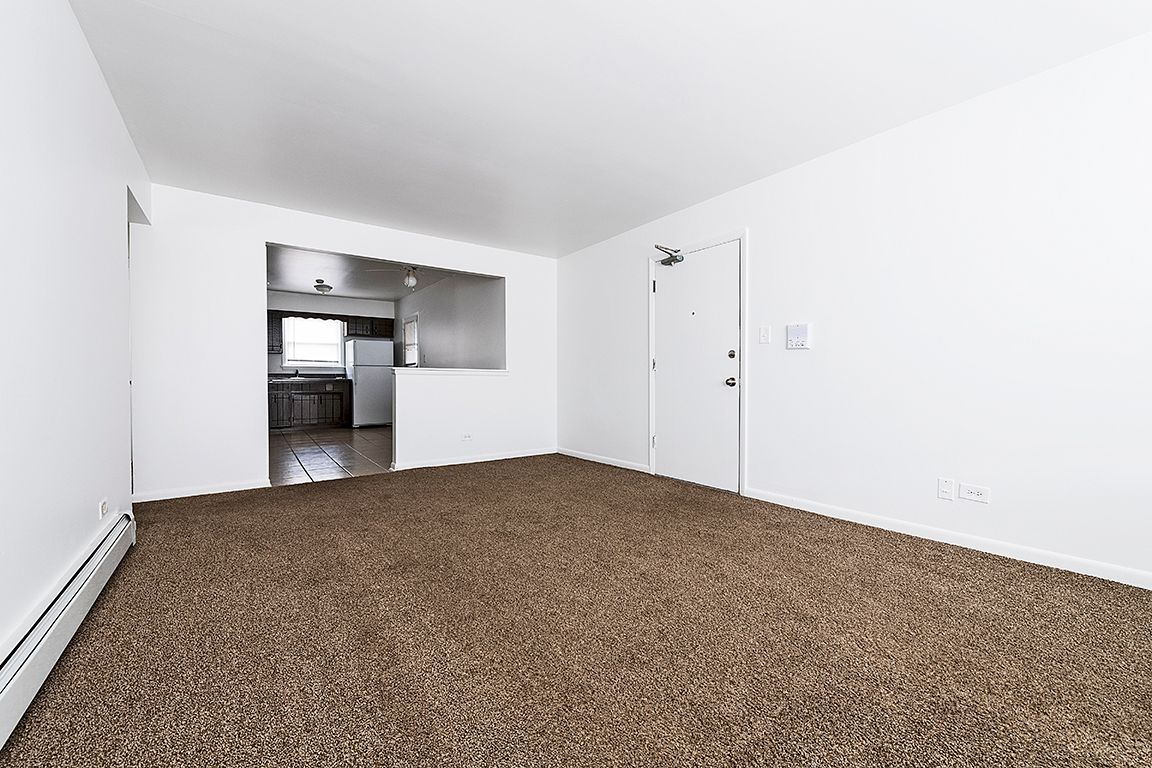 Empty living room with brown carpet, white walls, and a doorway to a kitchen.