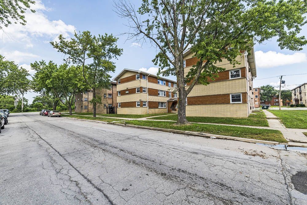 Apartment buildings on a cracked street. Trees line the sidewalk. Cars are parked.
