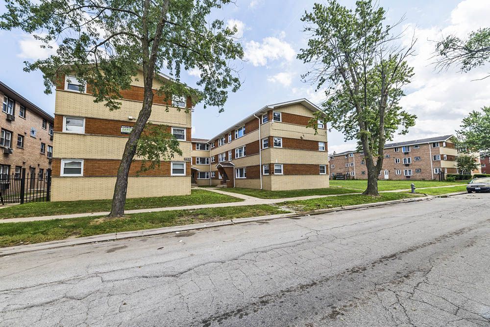 Apartment buildings with brick and yellow siding on a street with trees and cracked pavement.