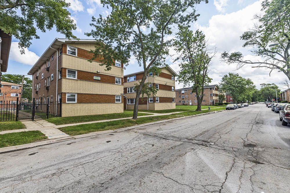 Apartment buildings line a cracked street with parked cars and trees under a sunny sky.