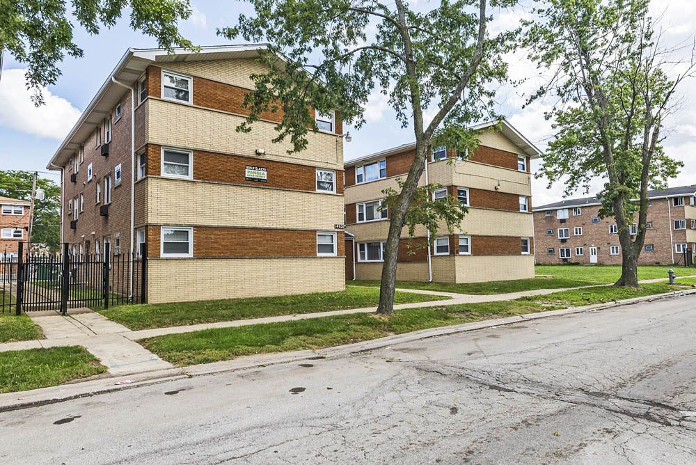 Two brick apartment buildings with light and dark brick accents, on a street with grass and trees.