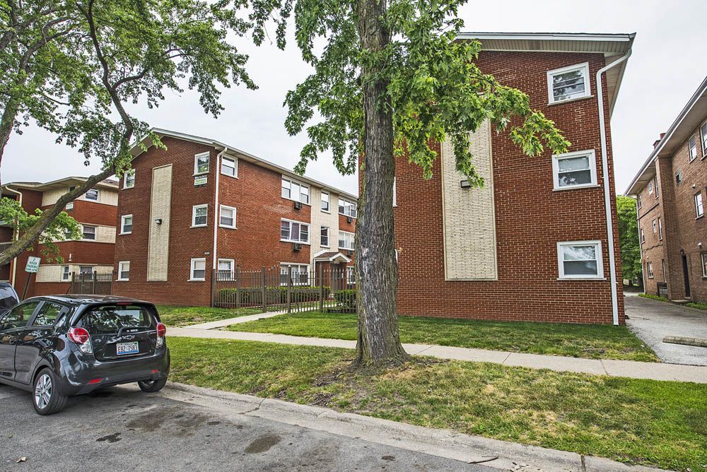 Brick apartment buildings with green lawn and trees. A black car parked on the street.