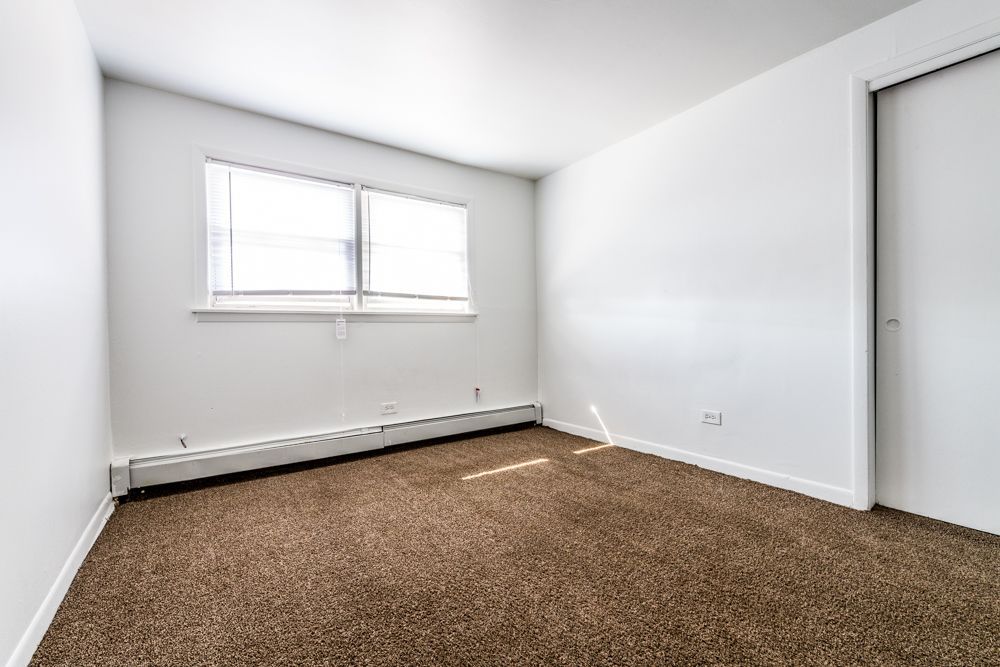 Empty bedroom with brown carpet, white walls, window, and closed closet door.