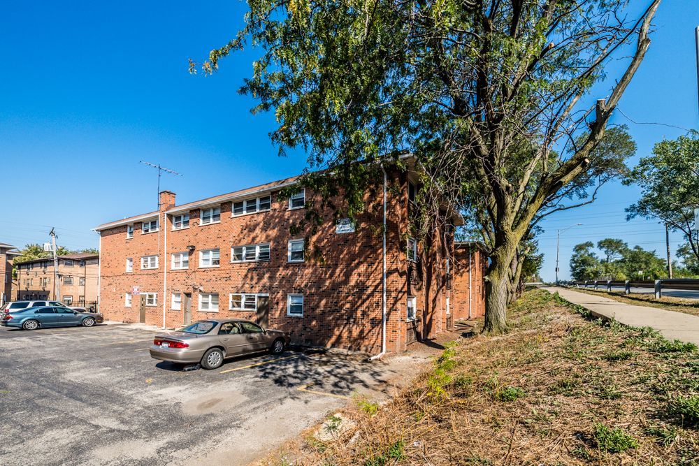 Brick apartment building with parked cars, trees, and blue sky.