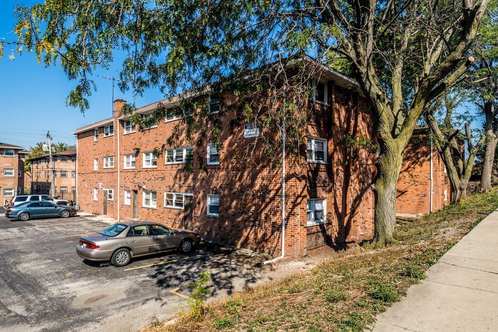 Red brick apartment building with cars parked in front, trees casting shadows.