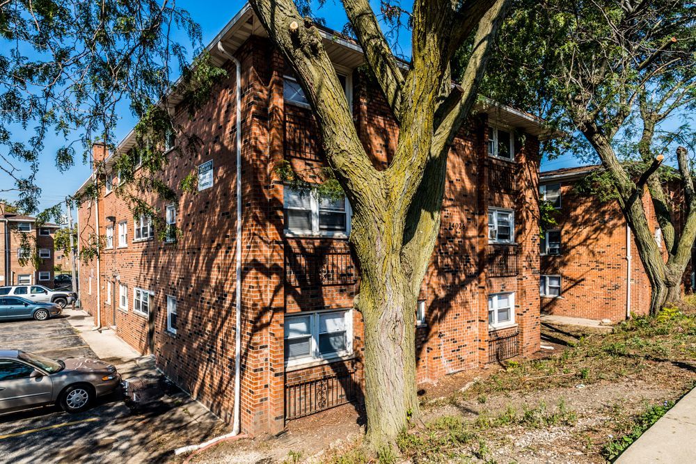 Red brick apartment buildings with white-framed windows, surrounded by trees and a parking lot.
