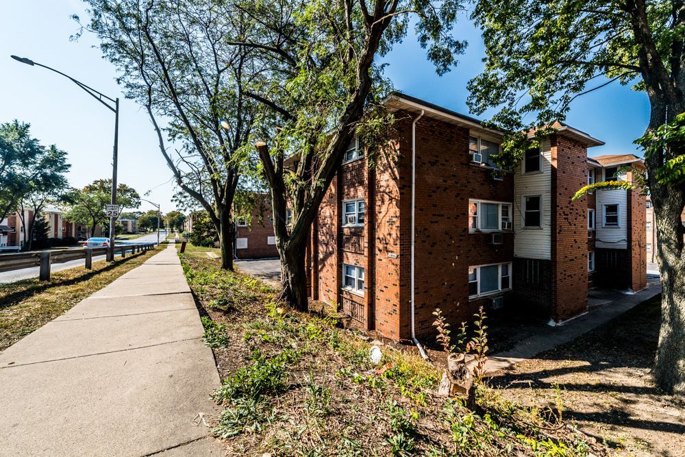 Brick apartment building next to a sidewalk with trees and a street.