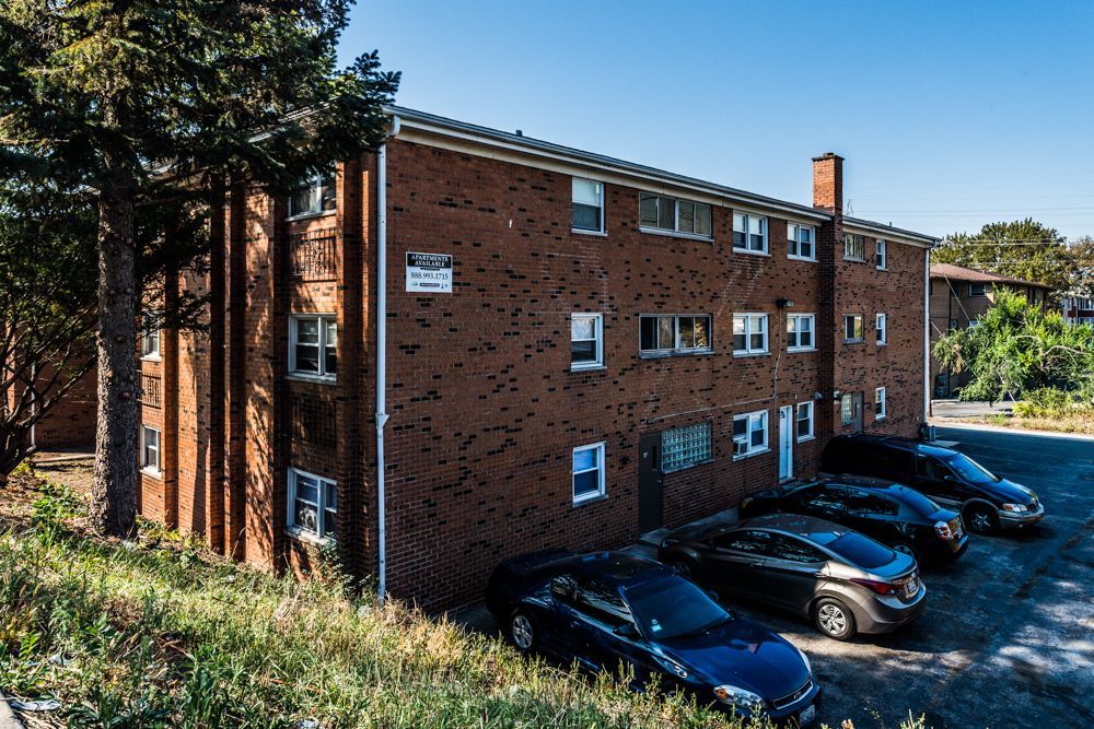 Brick apartment building with parked cars, surrounded by greenery.