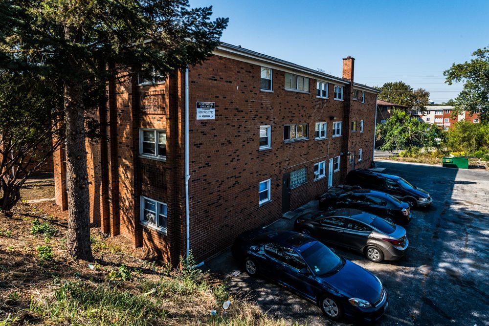 Three-story brick apartment building with parked cars, surrounded by trees and grass, on a sunny day.