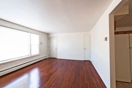 Empty room with wood floors, white walls, two doors, and a large window with sunlight.