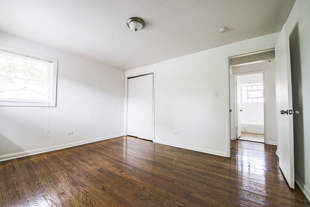 Empty bedroom with hardwood floors, white walls, closet, and open doorway to bathroom.
