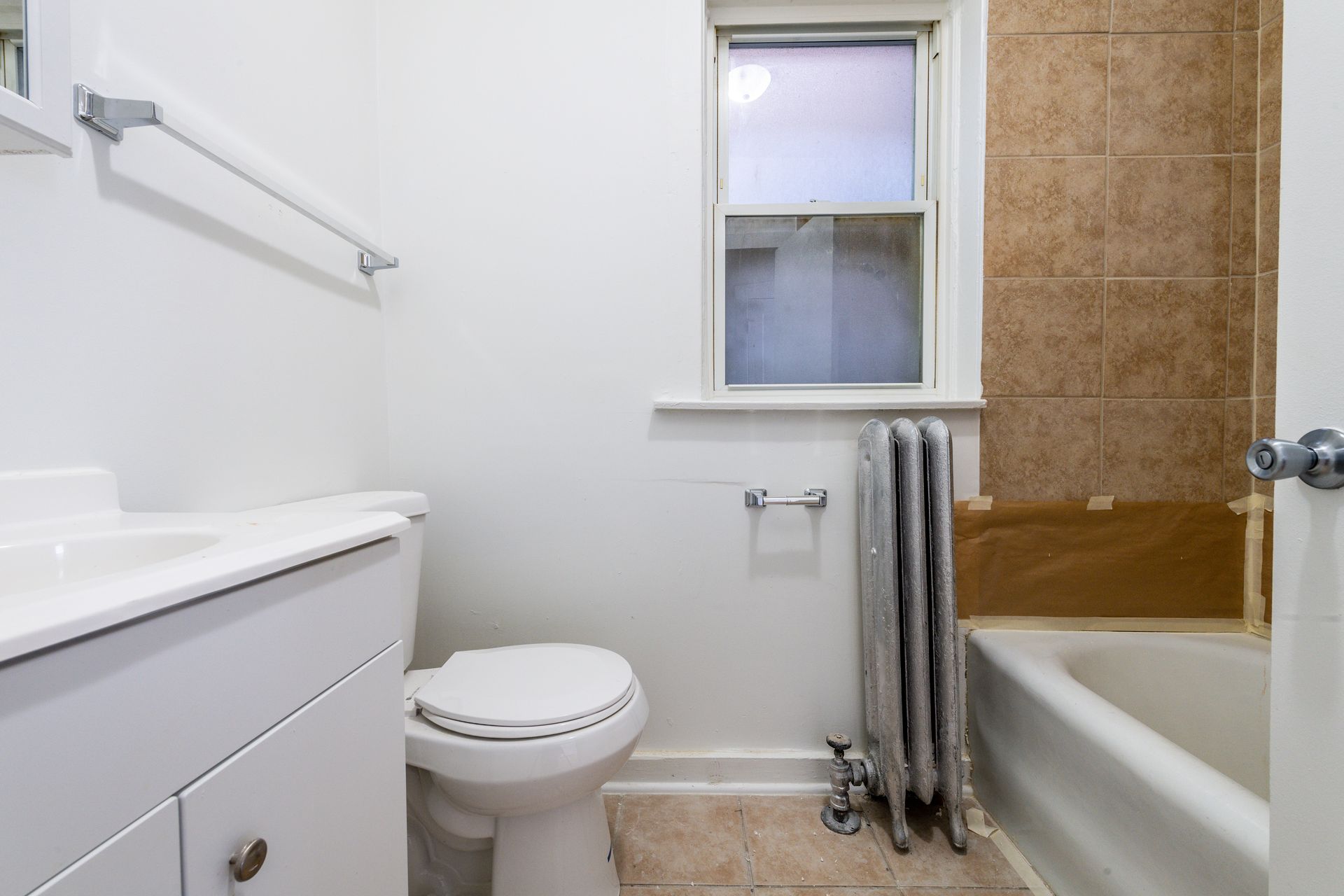 Bathroom with white vanity, toilet, and bathtub. Beige tiled wall, window, and radiator present.