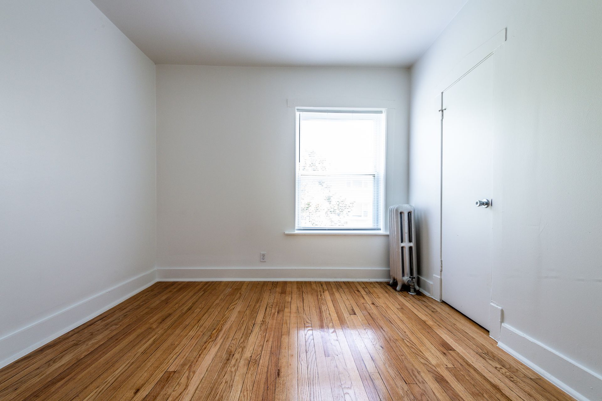 Empty room with wood floor, white walls, window, and door.