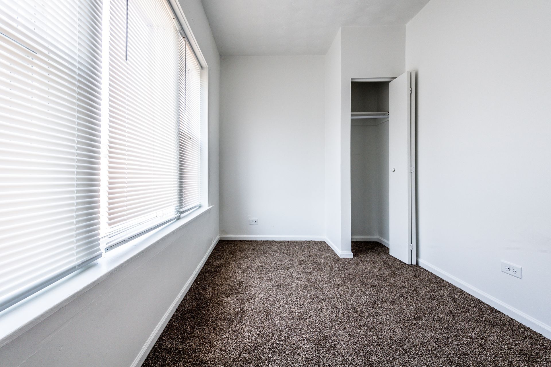 Empty room with white walls, brown carpet, window with blinds, and open closet.