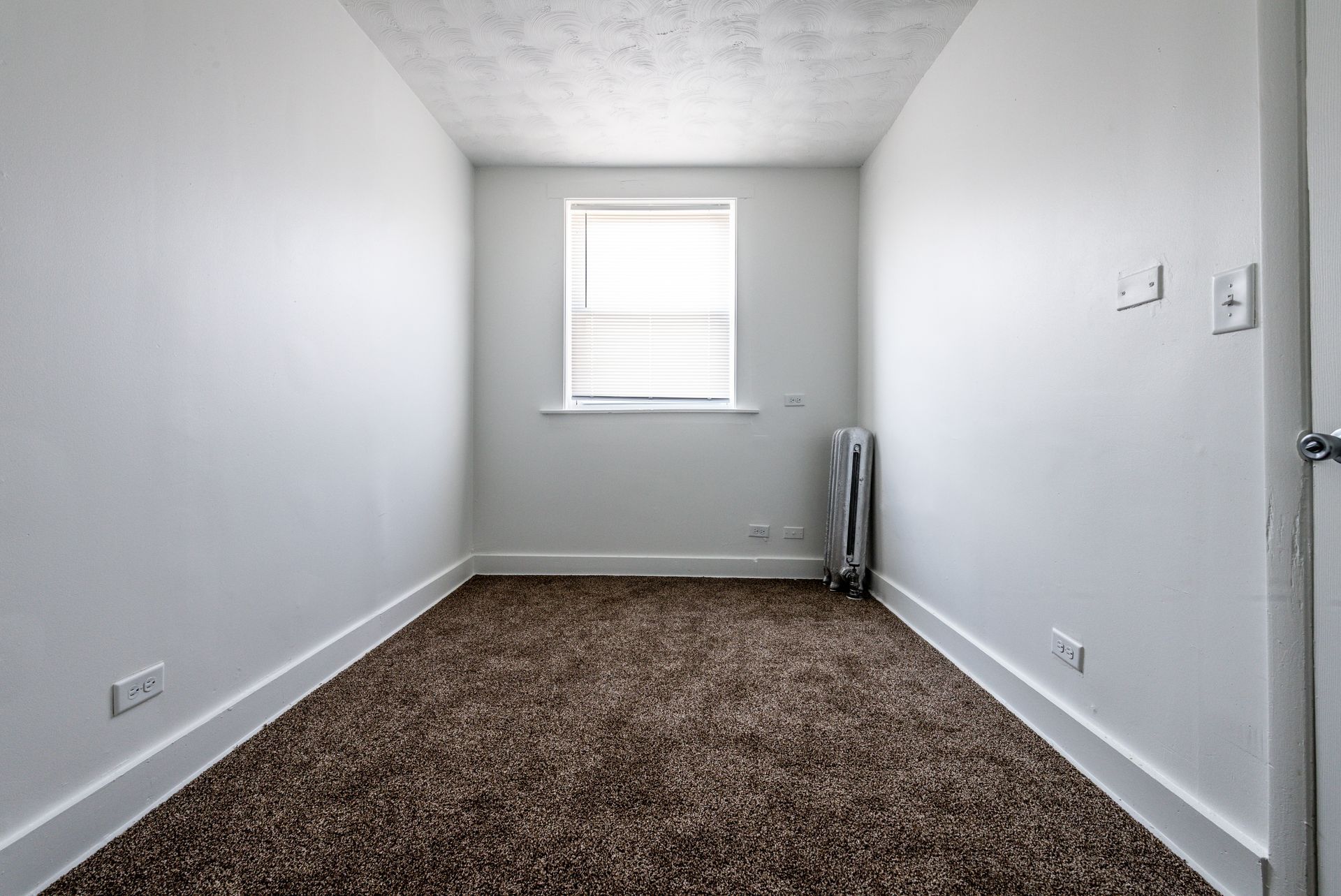 Empty, rectangular room with brown carpet, a window, and white walls and trim.